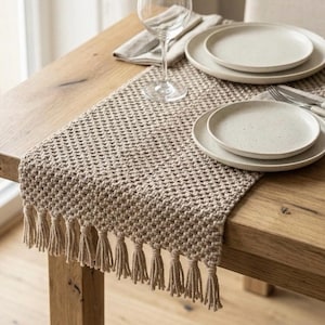 May include: A beige woven table runner with fringe detail, set on a wooden table. Two white plates, silverware, a wine glass, and a folded napkin are arranged on the runner, creating a neutral dining setting.