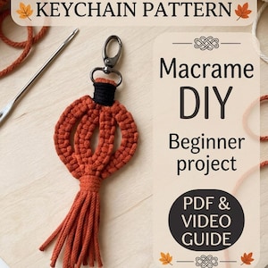 May include: A macrame pumpkin keyring with a black cord top and a metal lobster clasp. The text reads: HALLOWEEN PUMPKIN KEYCHAIN PATTERN, Macrame DIY Beginner project, PDF & VIDEO GUIDE, and Vuzlyk.Macrame. A metal crochet hook is visible nearby.
