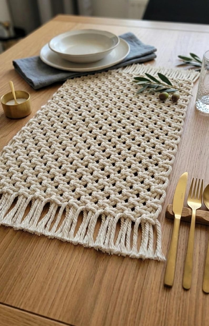 May include: A cream-colored macrame table runner with a fringed edge, set on a wooden table. The runner is rectangular and features a woven pattern. The table setting includes a white plate, bowl, and gold-colored cutlery.