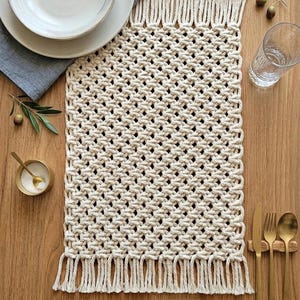 May include: A cream-colored macrame placemat with a fringed edge, set on a wooden table. The placemat is rectangular with a woven design. A white plate and bowl, a blue napkin, and gold cutlery are arranged on the table.