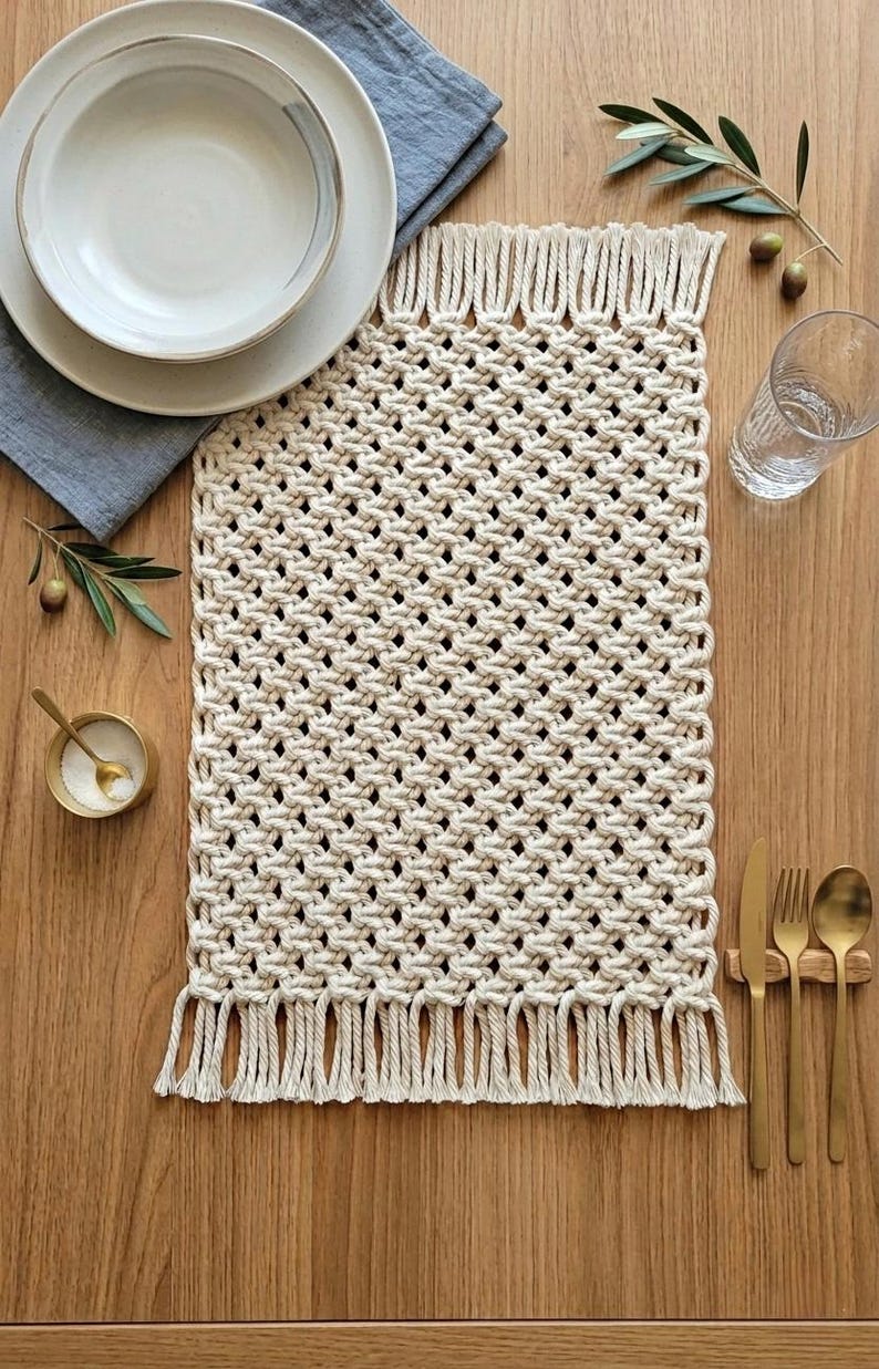 May include: A cream-colored macrame placemat with a fringed edge, set on a wooden table. The placemat is rectangular with a woven design. A white plate and bowl, a blue napkin, and gold cutlery are arranged on the table.
