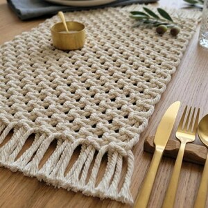May include: A beige macrame table runner with a fringed edge, set on a wooden table. Gold-colored flatware and a small bowl with a spoon are also present. The runner is approximately 12 inches wide.
