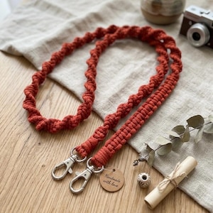 May include: A rust-colored macrame lanyard crafted with a spiral knot pattern. The lanyard features two silver-toned metal swivel lobster clasps at the ends and a small, round, light brown wooden tag with the text Handmade with love engraved on it.