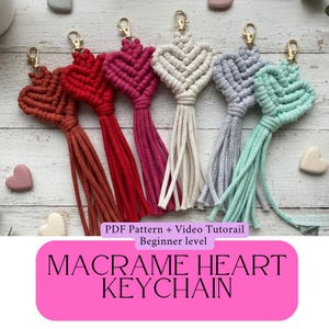 May include: A collection of macrame heart keychains in various colors, including red, pink, gray, and teal, each with a gold-colored clasp and a tassel. The keychains are handmade and feature a heart design. The text "MACRAME HEART KEYCHAIN" is displayed.