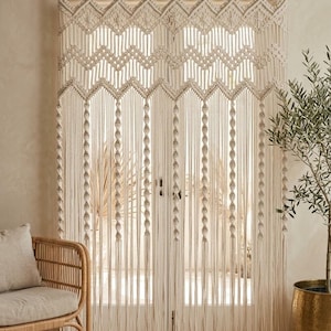 May include: An off-white macramé curtain featuring intricate knotted patterns, including diamond shapes and zig-zag lines at the top, with long vertical strands accented by small, evenly spaced spherical knots hanging from a dark metal rod.