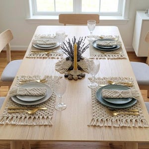 May include: A dining table set for four with macrame placemats, plates, gold-colored cutlery, and water glasses. A centerpiece includes a candle, lavender, and decorative stones. The table is made of light-colored wood.