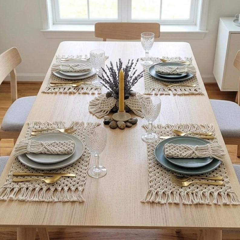 May include: A dining table set for four with macrame placemats, plates, gold-colored cutlery, and water glasses. A centerpiece includes a candle, lavender, and decorative stones. The table is made of light-colored wood.