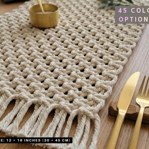 May include: A beige macrame table runner with fringe, measuring 12 x 18 inches. The text "45 COLOR OPTIONS" is visible. Gold-colored cutlery rests on a wooden surface, alongside a small gold bowl.