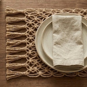 May include: A beige woven placemat with a macrame design and fringe detail. A white ceramic plate with a folded beige linen napkin is placed on top of the placemat. The table setting is on a wooden surface.