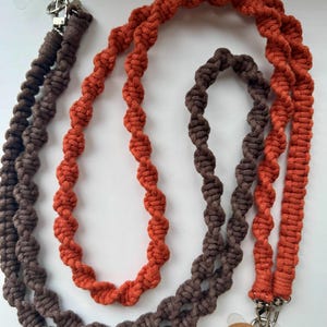 May include: Three macrame phone lanyards in shades of brown and orange. The lanyards have a spiral design and metal clasps. One lanyard has a small tag that reads "handmade with love".