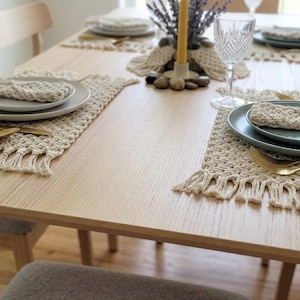 May include: A light wood dining table set for a meal. Each place setting includes a woven placemat with fringe, a plate, a napkin, and gold-colored cutlery. The table is decorated with a candle and a vase of dried flowers.