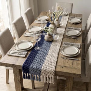 May include: A rectangular dining table with a wooden surface, set for six people with beige upholstered chairs. A textured table runner features a gradient of navy blue, light blue, and cream, finished with fringe tassels at both ends.