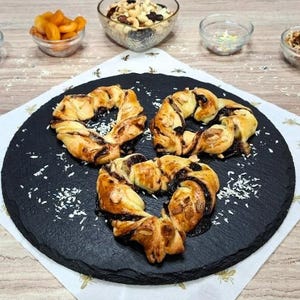 May include: Three heart-shaped pastries with a dark filling and almond slices on a black slate plate. The pastries are golden brown with a glossy finish. Small bowls of dried apricots, nuts, and sprinkles surround the plate, suggesting a bakery or dessert theme.