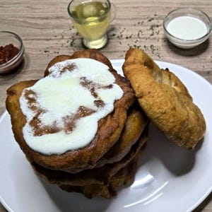 May include: A stack of golden-brown fried dough, topped with a creamy white sauce, is on a white plate. A separate piece of fried dough is beside the stack. Small bowls of red sauce, oil, and white sauce are in the background.