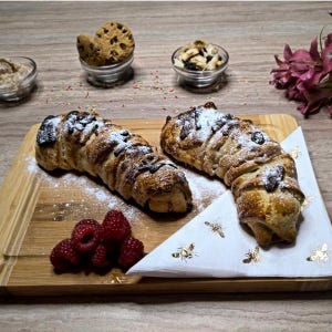 Peut inclure: Deux pâtisseries saupoudrées de sucre glace et nappées de chocolat sont présentées sur une planche en bois. Des framboises fraîches et des petits bols de biscuits et de noix complètent la scène, évoquant un dessert.
