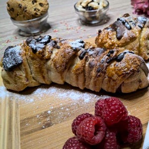 Peut inclure: Gros plan sur une planche en bois avec des pâtisseries et des framboises fraîches. Les pâtisseries comprennent une longue pâtisserie torsadée avec du chocolat et du sucre glace, et deux plus petites pâtisseries. Des bols en verre contiennent des biscuits et des noix.