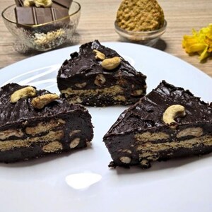 Può includere: Tre fette triangolari di dessert al cioccolato su un piatto bianco. Il dessert ha una copertura di cioccolato fondente con anacardi. La base sembra essere fatta di biscotti. Cioccolato e noci aggiuntivi sullo sfondo.