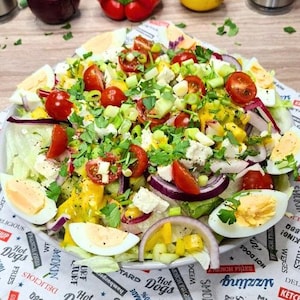 May include: A colorful salad on a white plate, featuring halved hard-boiled eggs, red cherry tomatoes, red onion slices, and green lettuce. The salad is garnished with fresh herbs and a yellow dressing. The plate rests on a paper with text.