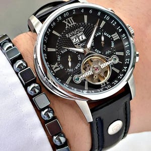 Puede incluir: Un reloj de pulsera Raymond negro y plateado con una correa de cuero negro. La esfera muestra la fecha, el día de la semana y el mes. También se ve una pulsera negra y plateada a juego.