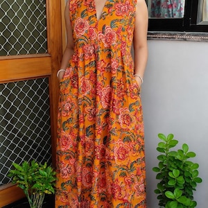 May include: A sleeveless floor-length dress in a vibrant orange hue featuring a repeating floral pattern in shades of red, pink, and dark green. The garment has a V-neckline with a collar, a fitted bodice, and a full-length skirt with side pockets.