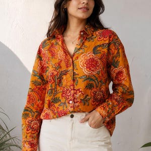 May include: A long-sleeved button-down shirt in a vibrant mustard yellow hue, featuring a detailed floral pattern in shades of red, orange, and dark green. The shirt has a classic collar, a front button placket, and is paired with off-white trousers.