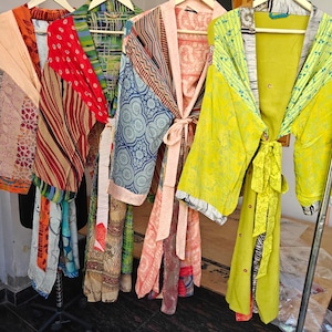 May include: A collection of colorful, patterned kimonos hanging on a clothing rack. The kimonos feature various designs, including floral, geometric, and abstract patterns in shades of orange, red, blue, and green. Each kimono has a tie closure.