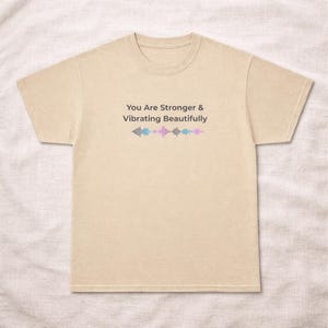 Pode incluir: Uma camiseta bege com o texto "You Are Stronger & Vibrating Beautifully" em preto. Abaixo do texto, um gráfico de onda sonora em cinza, azul e rosa. A camiseta tem mangas curtas e gola redonda.