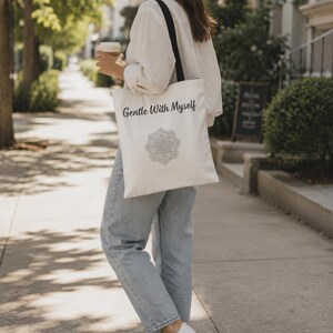K&ouml;nnte beinhalten: Cremefarbene Tragetasche mit schwarzen Tr&auml;gern. Auf der Tasche steht der Text "Gentle With Myself" &uuml;ber einem Mandala-Design. Die Tasche wird von einer Person getragen, die eine hellblaue Jeans und ein wei&szlig;es Hemd tr&auml;gt.
