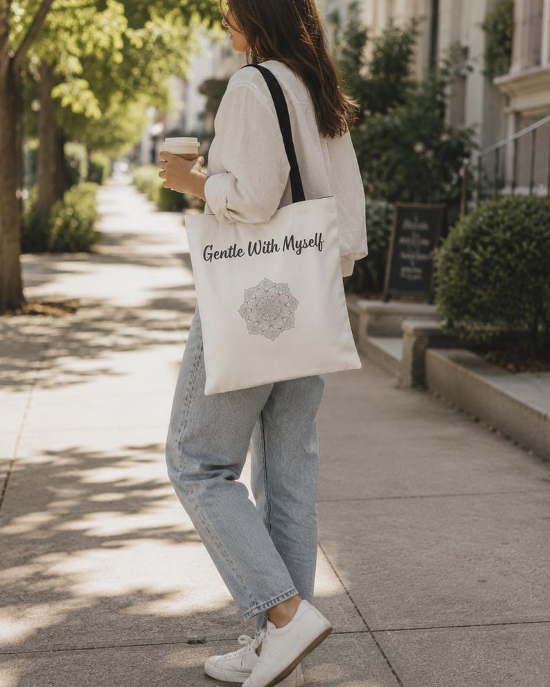 K&ouml;nnte beinhalten: Cremefarbene Tragetasche mit schwarzen Tr&auml;gern. Auf der Tasche steht der Text "Gentle With Myself" &uuml;ber einem Mandala-Design. Die Tasche wird von einer Person getragen, die eine hellblaue Jeans und ein wei&szlig;es Hemd tr&auml;gt.