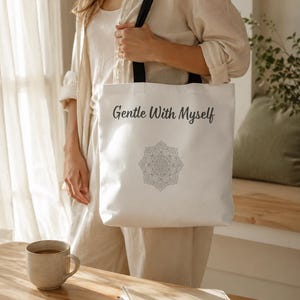 K&ouml;nnte beinhalten: Wei&szlig;er Shopper mit schwarzen Tr&auml;gern, mit dem Text "Gentle With Myself" und einem Mandala-Design. Die Tasche wird von einer Person in einem beigen Outfit gehalten. Eine Tasse und ein Buch liegen auf einem Holztisch.