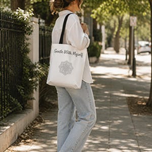 K&ouml;nnte beinhalten: Eine wei&szlig;e Tragetasche mit schwarzen Tr&auml;gern. Auf der Tasche steht "Gentle With Myself" und ein schwarz-wei&szlig;es Mandala-Design. Die Tasche wird von einer Person getragen, die ein wei&szlig;es Hemd und eine blaue Jeans tr&auml;gt.