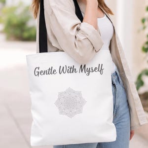 K&ouml;nnte beinhalten: Wei&szlig;e Tragetasche mit schwarzen Tr&auml;gern. Auf der Tasche steht der Text "Gentle With Myself" in Schreibschrift, darunter ein graues Mandala-Design. Die Tasche wird &uuml;ber der Schulter getragen.