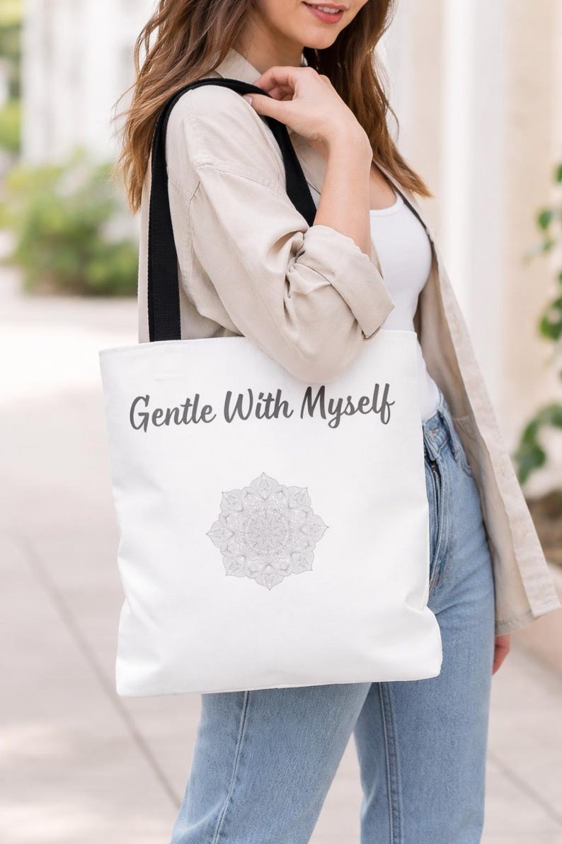 K&ouml;nnte beinhalten: Wei&szlig;e Tragetasche mit schwarzen Tr&auml;gern. Auf der Tasche steht der Text "Gentle With Myself" in Schreibschrift, darunter ein graues Mandala-Design. Die Tasche wird &uuml;ber der Schulter getragen.