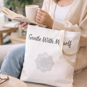K&ouml;nnte beinhalten: Eine wei&szlig;e Tragetasche mit dem Text "Gentle With Myself" in schwarzer Schrift und einem grauen Mandala-Design. Die Tasche hat beige Tr&auml;ger und wird von einer Person gehalten. Die Person h&auml;lt ein Buch und eine Tasse.