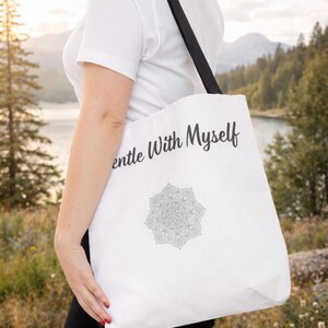 K&ouml;nnte beinhalten: Wei&szlig;e Tragetasche mit schwarzen Henkeln. Auf der Tasche steht der Text "Gentle With Myself" und ein Mandala-Design. Der Hintergrund zeigt einen See und Berge. Die Tasche wird &uuml;ber der Schulter getragen.