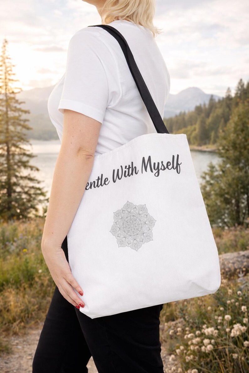 K&ouml;nnte beinhalten: Wei&szlig;e Tragetasche mit schwarzen Henkeln. Auf der Tasche steht der Text "Gentle With Myself" und ein Mandala-Design. Der Hintergrund zeigt einen See und Berge. Die Tasche wird &uuml;ber der Schulter getragen.