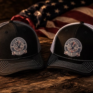Puede incluir: Dos gorras de béisbol con paneles frontales negros y paneles traseros de malla blanca. Cada gorra presenta un parche bordado circular que muestra un águila, la bandera estadounidense y el texto 250TH YEARS OF INDEPENDENCE 1776-2026 al frente.