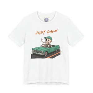 Puede incluir: Camiseta blanca con un dibujo animado de un esqueleto conduciendo un descapotable turquesa. El esqueleto lleva una gorra verde y fuma. Las palabras "JUST CALM" están escritas en naranja encima del coche.