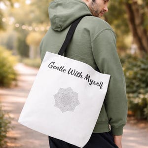 K&ouml;nnte beinhalten: Wei&szlig;e Tragetasche mit dem Text "Gentle With Myself" und einem Mandala-Design. Die Tasche hat einen schwarzen Riemen und wird &uuml;ber der Schulter getragen. Der Hintergrund ist eine verschwommene Au&szlig;enszene.