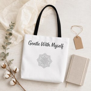 K&ouml;nnte beinhalten: Wei&szlig;e Tragetasche mit schwarzen Henkeln, mit dem Text "Gentle With Myself" und einem grauen Mandala-Design. Auf einer wei&szlig;en Oberfl&auml;che liegen Baumwollzweige und ein kleines Notizbuch.