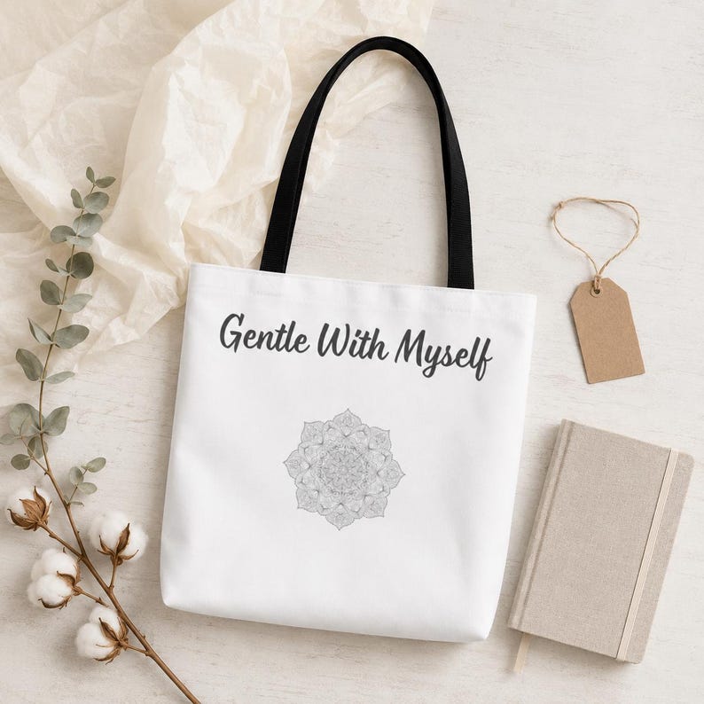 K&ouml;nnte beinhalten: Wei&szlig;e Tragetasche mit schwarzen Henkeln, mit dem Text "Gentle With Myself" und einem grauen Mandala-Design. Auf einer wei&szlig;en Oberfl&auml;che liegen Baumwollzweige und ein kleines Notizbuch.