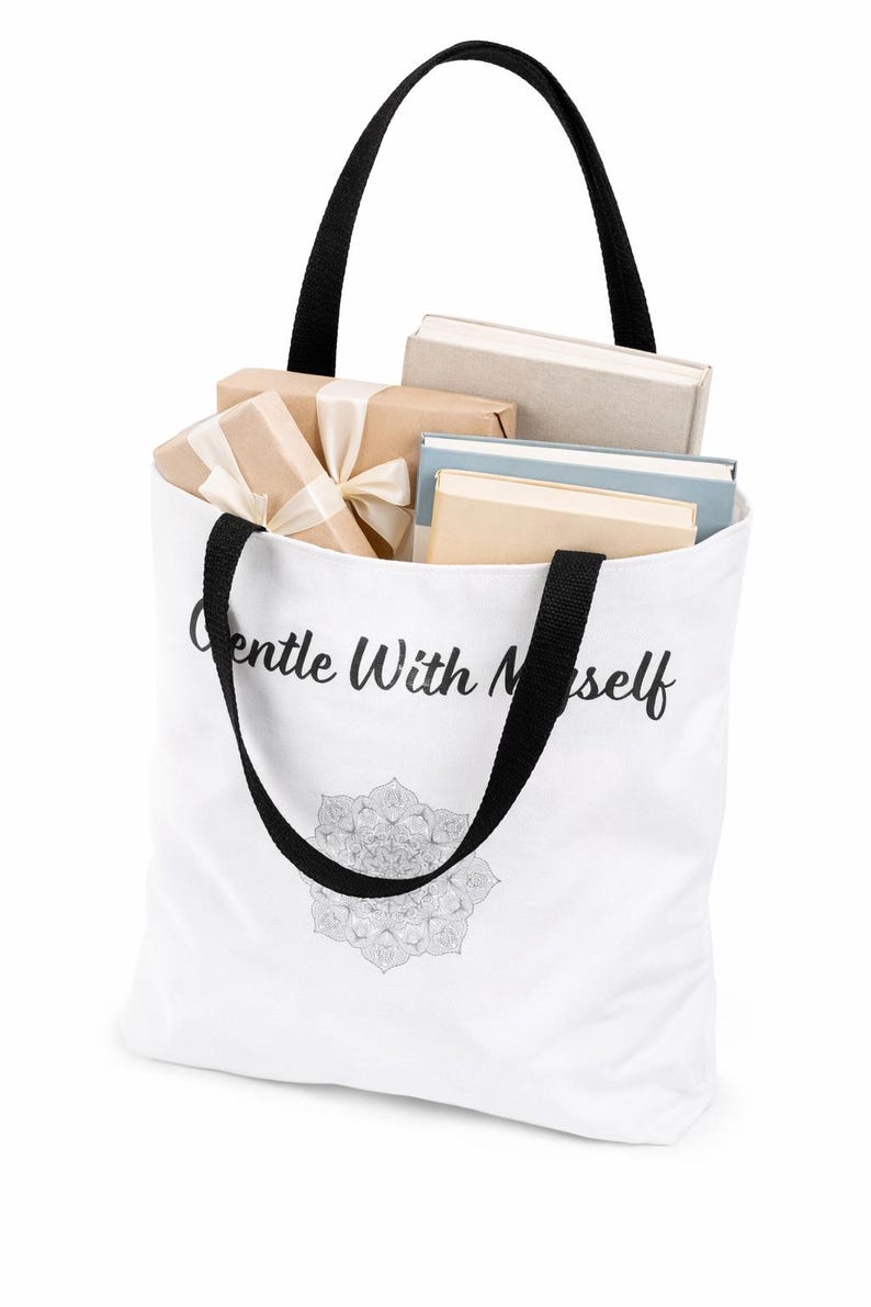 K&ouml;nnte beinhalten: Wei&szlig;e Tragetasche mit schwarzen Henkeln, gef&uuml;llt mit B&uuml;chern und verpackten Geschenken. Die Tasche tr&auml;gt den Schriftzug "Gentle With Myself" in Schreibschrift sowie ein detailliertes Mandala-Design am unteren Rand.