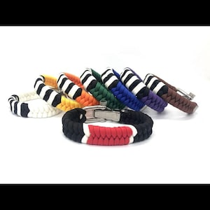 Op de afbeelding: Een verzameling kleurrijke paracord-armbanden met roestvrijstalen sluitingen. De armbanden hebben verschillende kleurencombinaties, waaronder zwart, rood, wit en gestreepte patronen. De armbanden worden op een wit oppervlak gepresenteerd.