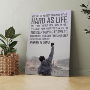 May include: A rectangular wall art print featuring a person in a grey hooded sweatshirt with one arm raised in the air. The background shows a snowy park scene. The text reads: YOU, ME, OR NOBODY IS GONNA HIT AS HARD AS LIFE. THAT'S HOW WINNING IS DONE!