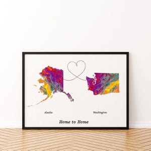 May include: A framed wall art print featuring a colourful, abstract paint-swirl map of Alaska and Washington connected by a thin line forming a heart shape. The text Alaska and Washington appear below the states, with Home to Home at the bottom.