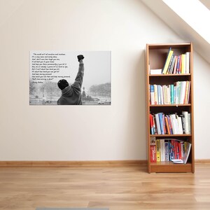 May include: Black and white framed print featuring a motivational quote and an image of a person with their fist raised in the air. The print is displayed on a white wall, with a wooden bookshelf to the right.
