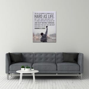 May include: A rectangular wall art print featuring a person in a grey hooded sweatshirt standing with one arm raised in a snowy outdoor setting. The text reads: YOU, ME, OR NOBODY IS GONNA HIT AS HARD AS LIFE. BUT IT AIN'T ABOUT HOW HARD YA HIT.