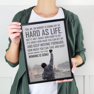 May include: A black framed wall art print featuring a gray-toned photograph of a person in a hoodie with one arm raised in victory. The text reads: YOU, ME, OR NOBODY IS GONNA HIT AS HARD AS LIFE. BUT IT AINT ABOUT HOW HARD YA HIT. IT IS ABOUT HOW HARD YOU CAN GET HIT AND KEEP MOVING FORWARD. HOW MUCH YOU CAN TAKE AND KEEP MOVING FORWARD. THAT IS HOW WINNING IS DONE!