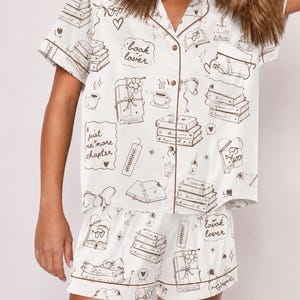 May include: A white short-sleeve button-down pajama shirt and matching shorts set featuring a brown line-art print of stacks of books, coffee cups, and text that reads "book lover" and "just one more chapter" with brown piping trim.