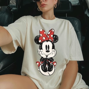 May include: Cream-colored t-shirt featuring a graphic of Minnie Mouse. Minnie is depicted in black and white with a red polka-dot bow and matching skirt. The shirt has short sleeves and a relaxed fit, suitable for casual wear.